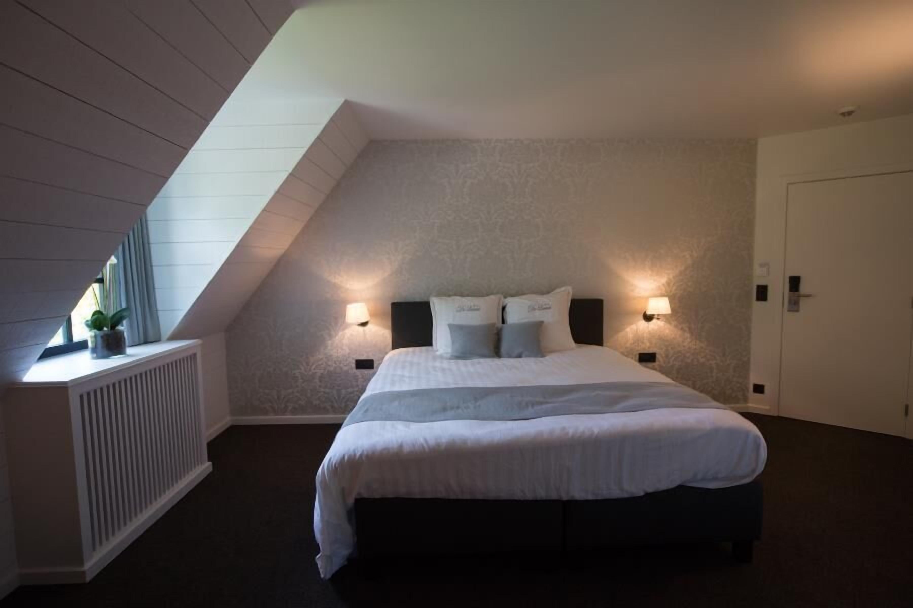 A bedroom with a sloped ceiling, a bed with white linens, two bedside lamps, and a radiator.