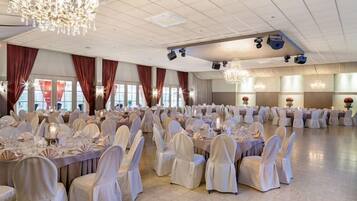 A banquet hall with round tables, white chairs, chandeliers, and large windows.