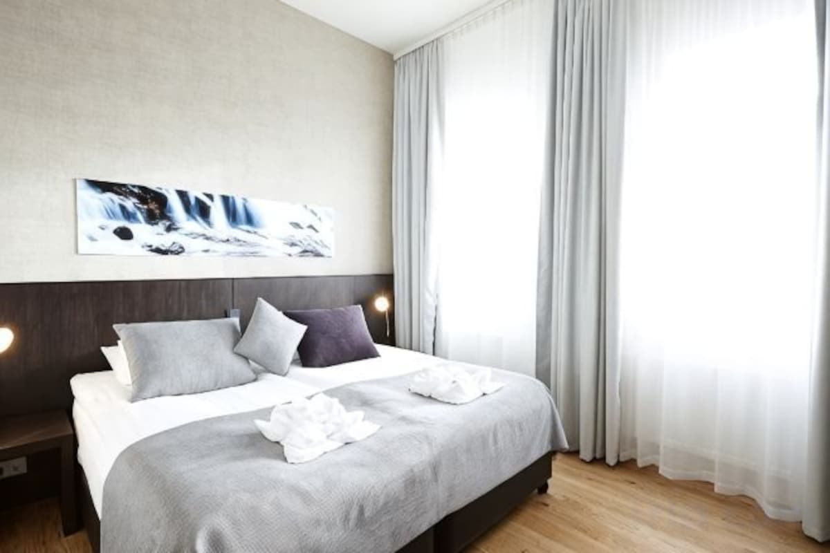 A modern hotel room with a large bed, a headboard, a wall-mounted artwork, and a window with curtains.