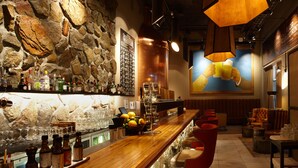 Zona de bar con pared de piedra, mostrador de madera y varias botellas. Hay taburetes y una zona de estar con mesa y sillas.