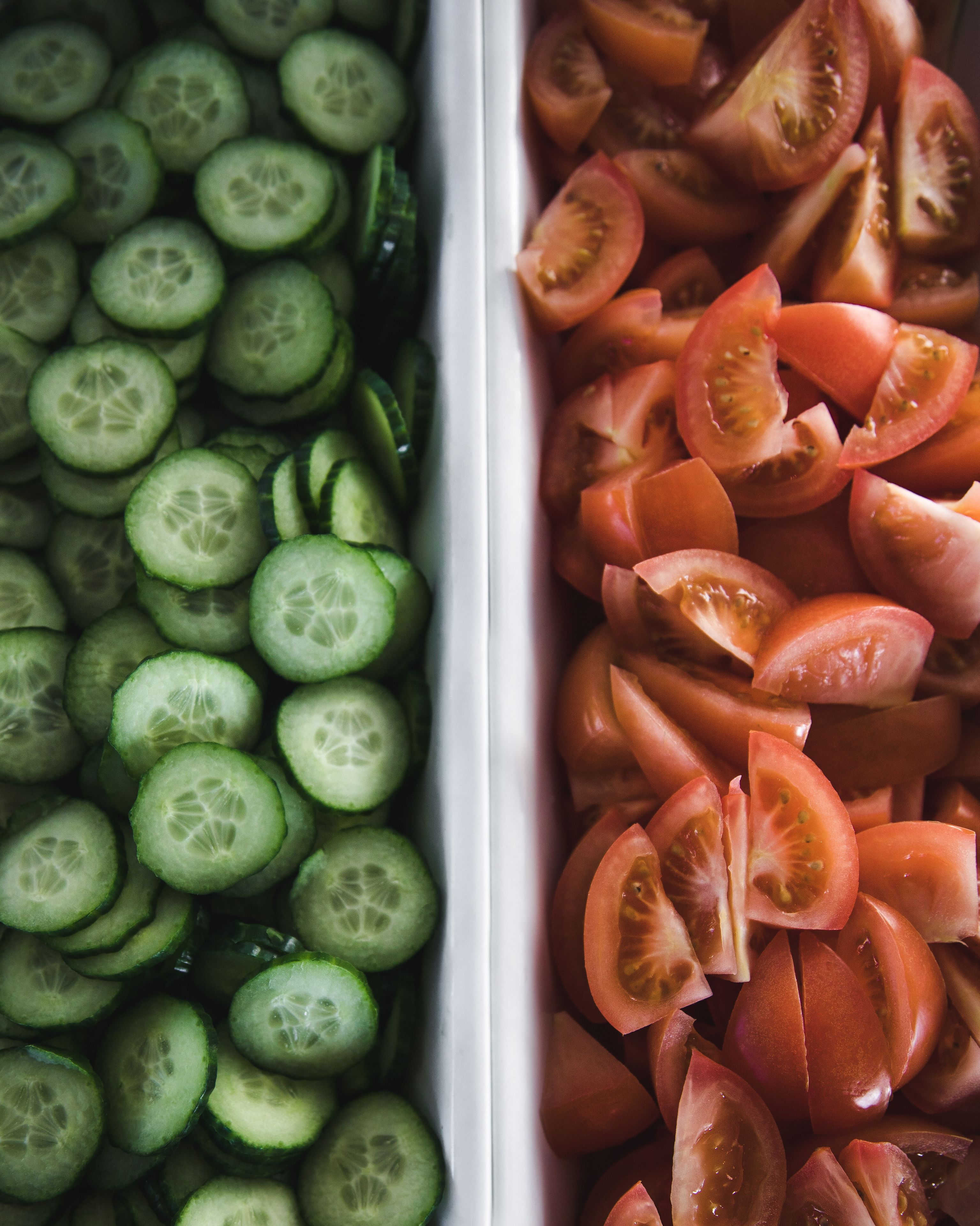 A white tray divided into two sections, one filled with sliced cucumbers and the other with sliced tomatoes.
