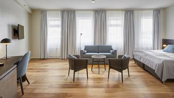 Quarto de hotel moderno com sofá, duas poltronas, uma mesa pequena, cama e escrivaninha com luminária.