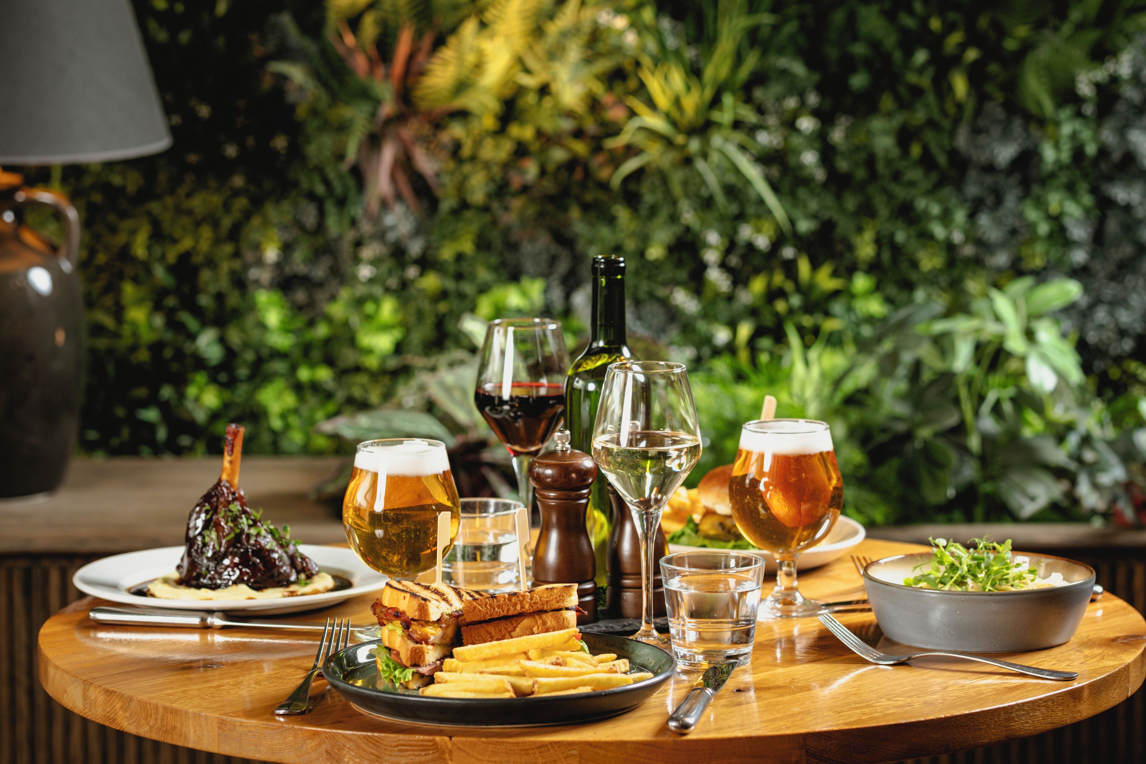 Mesa de madeira redonda com uma refeição, copos de cerveja e uma garrafa de vinho, em frente a um fundo verde exuberante.