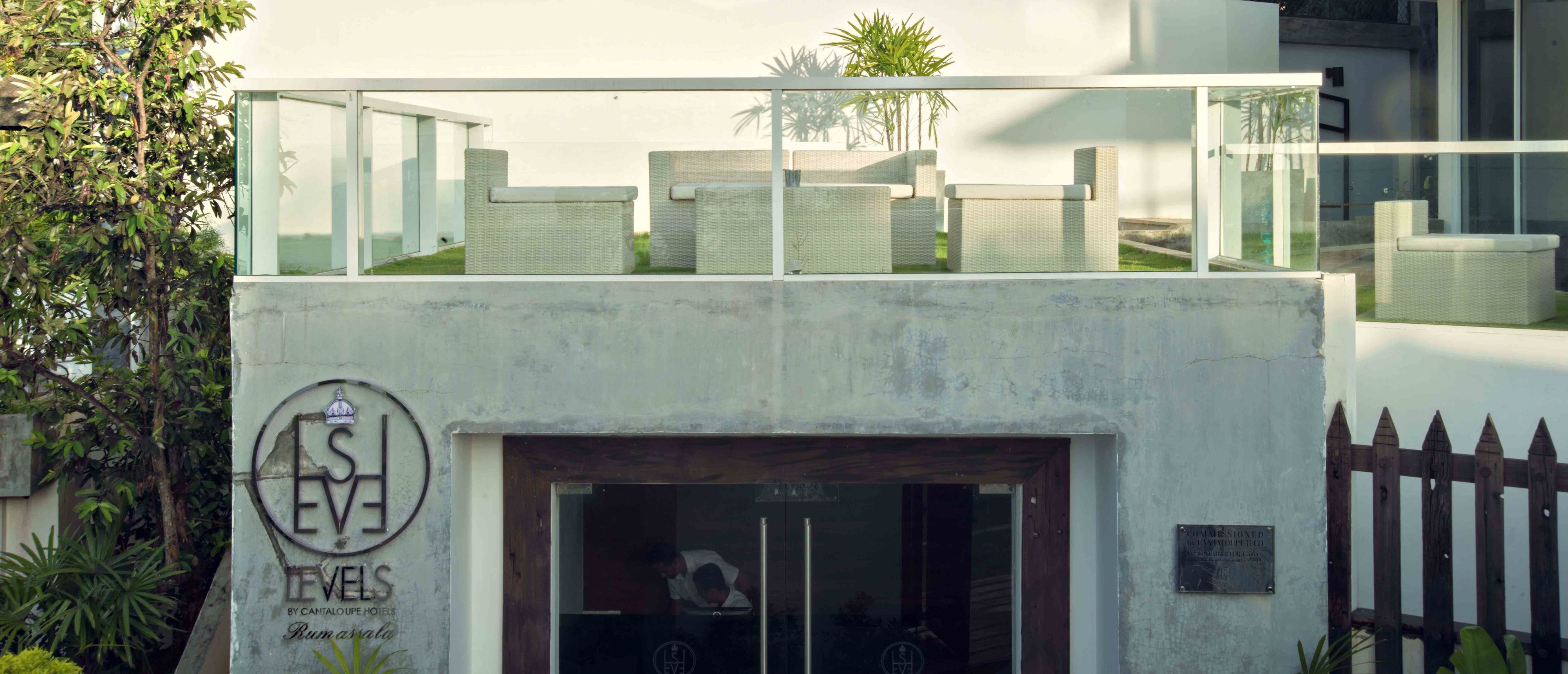 A hotel entrance with a glass door, a signboard, and a view of outdoor seating and greenery.