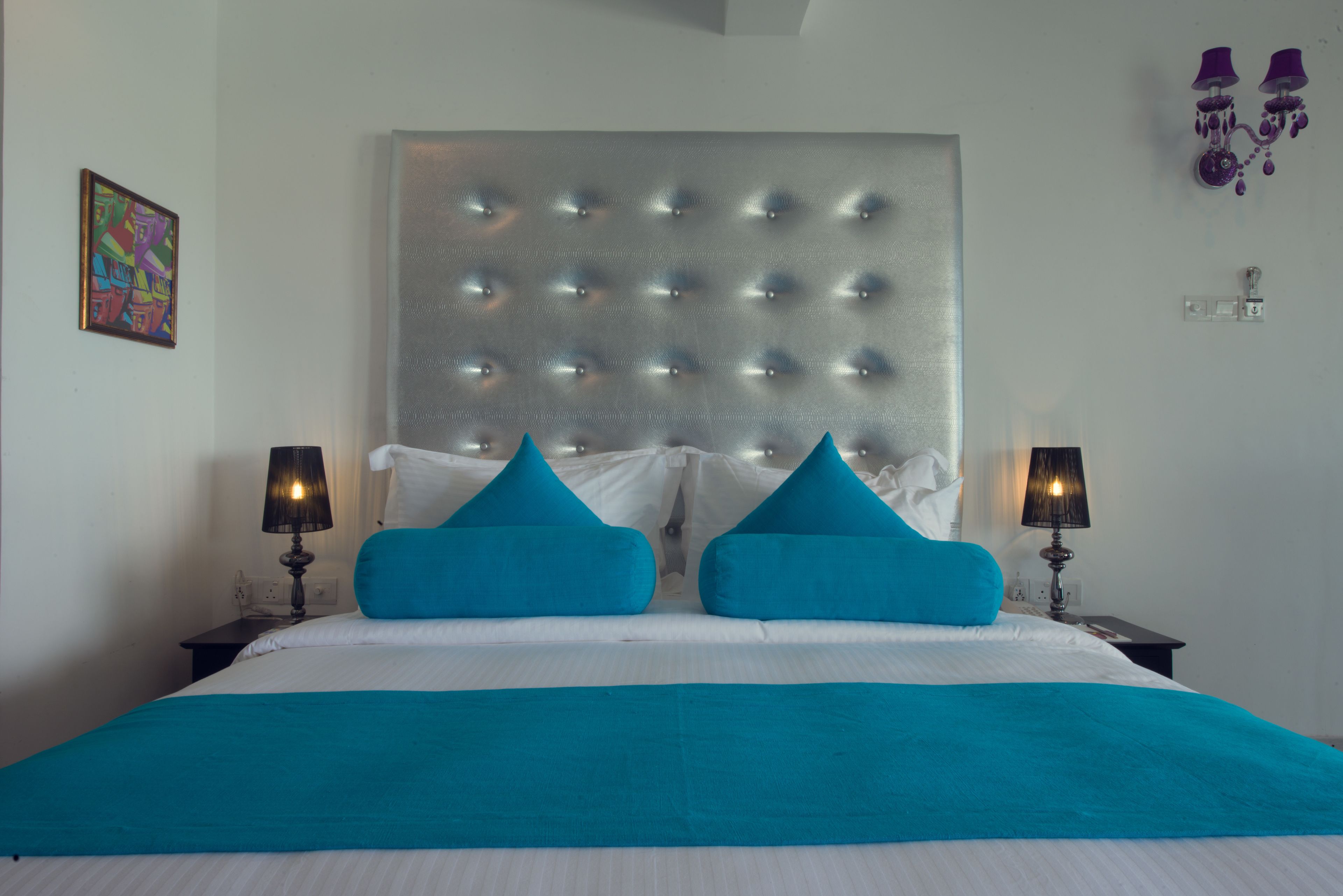 Een netjes opgemaakt bed met een gestuufd hoofdbord, twee nachtkastjes met lampen, en een kleurrijk schilderij aan de muur.