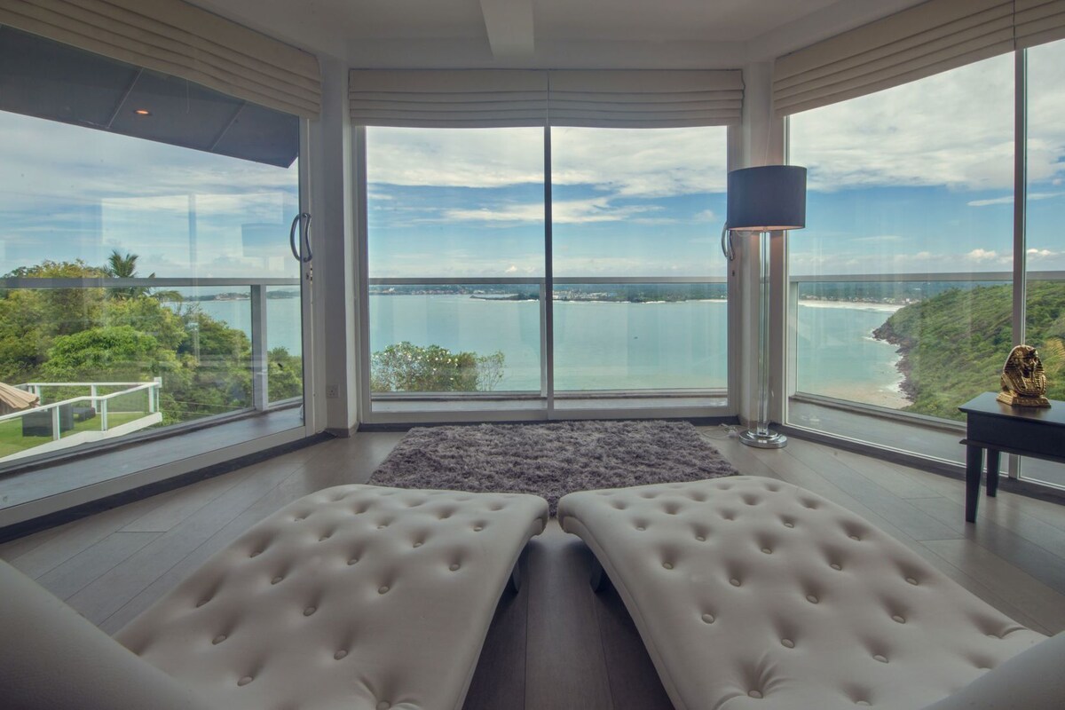 Une chambre moderne dotée de grandes fenêtres, de deux coins salon avec des fauteuils confortables, d’un lampadaire et offrant une vue sur une étendue d’eau.