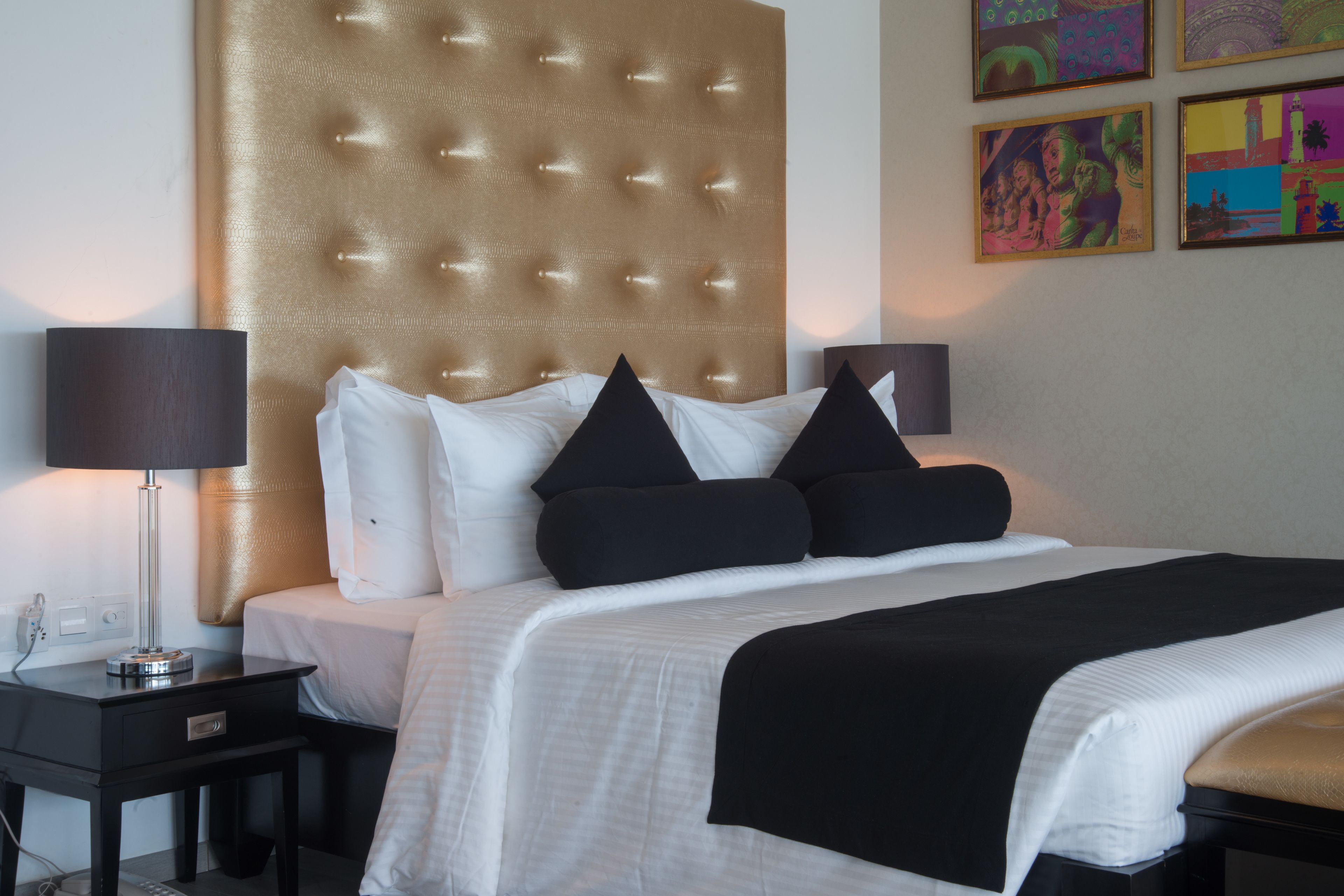 A hotel room with a tufted headboard, a bed with black and white bedding, a bedside table with a lamp, and artwork on the wall.