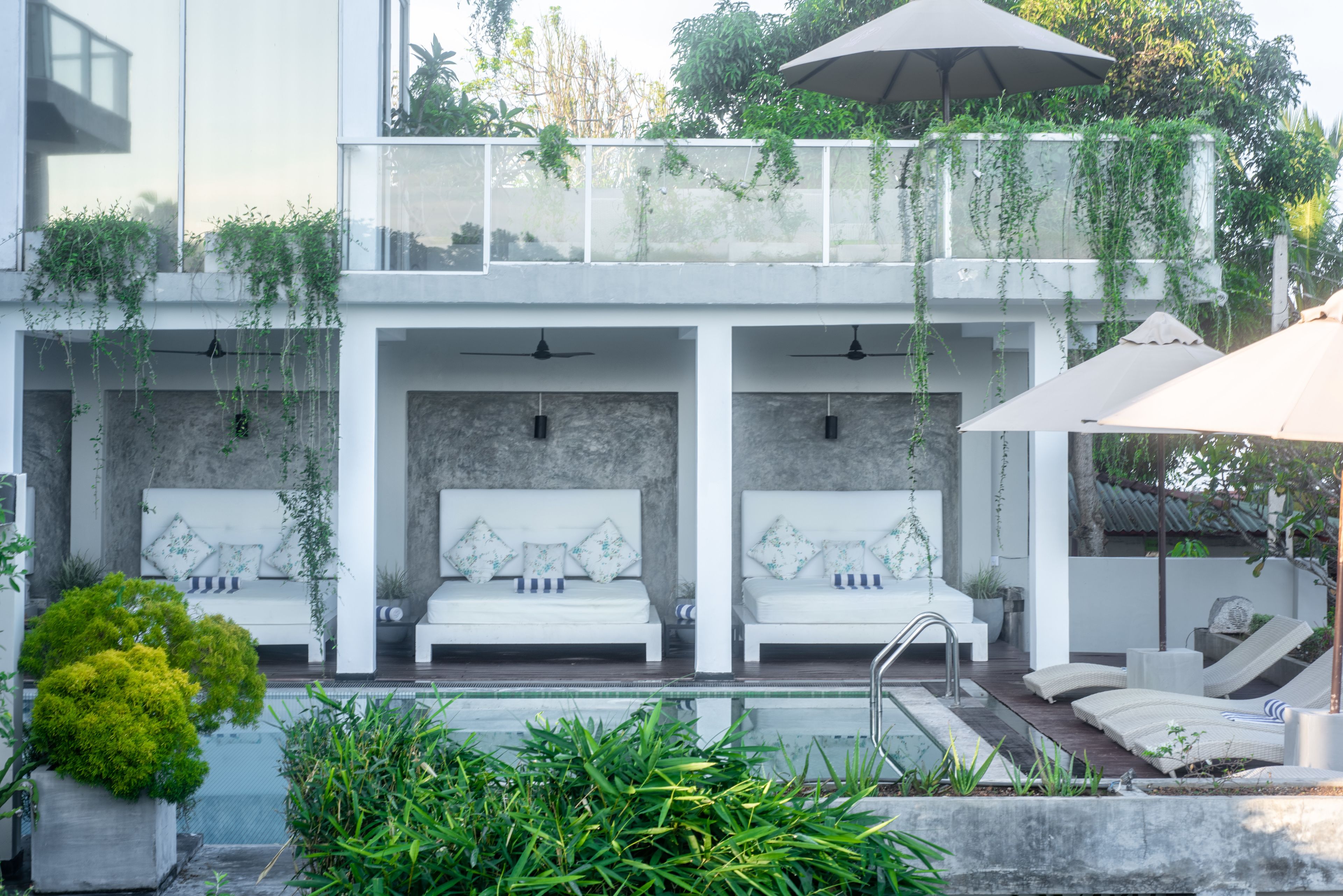 Uma área de estar moderna ao ar livre, com sofás brancos e guarda-sóis, cercada por vegetação e uma piscina.