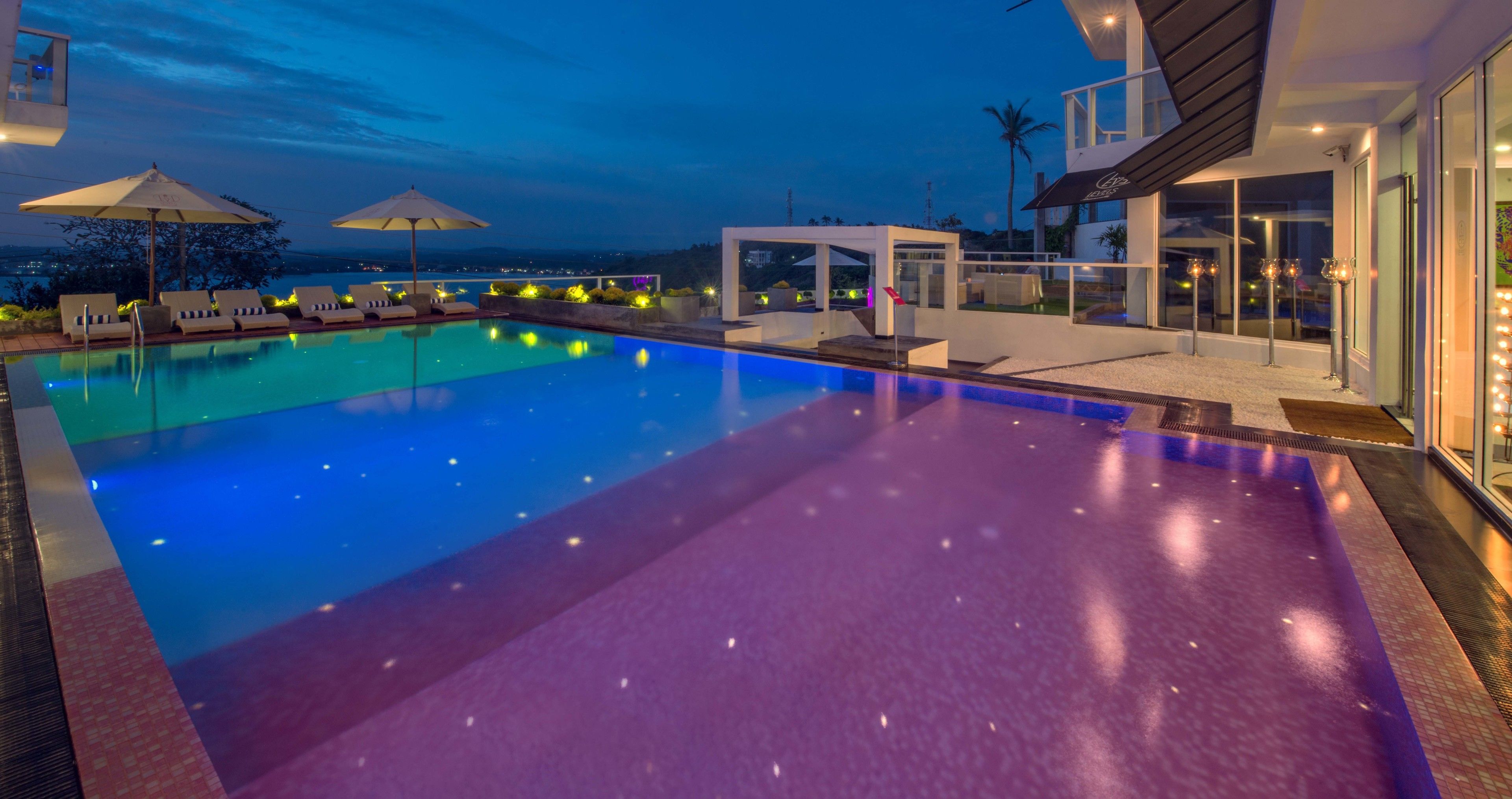 A swimming pool with lounge chairs and umbrellas, a covered seating area, and a view of the city at dusk.