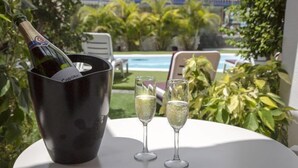 Een fles champagne in een zwarte emmer met twee glazen op een witte tafel, buiten gezet met een zwembad en groen in de achtergrond.
