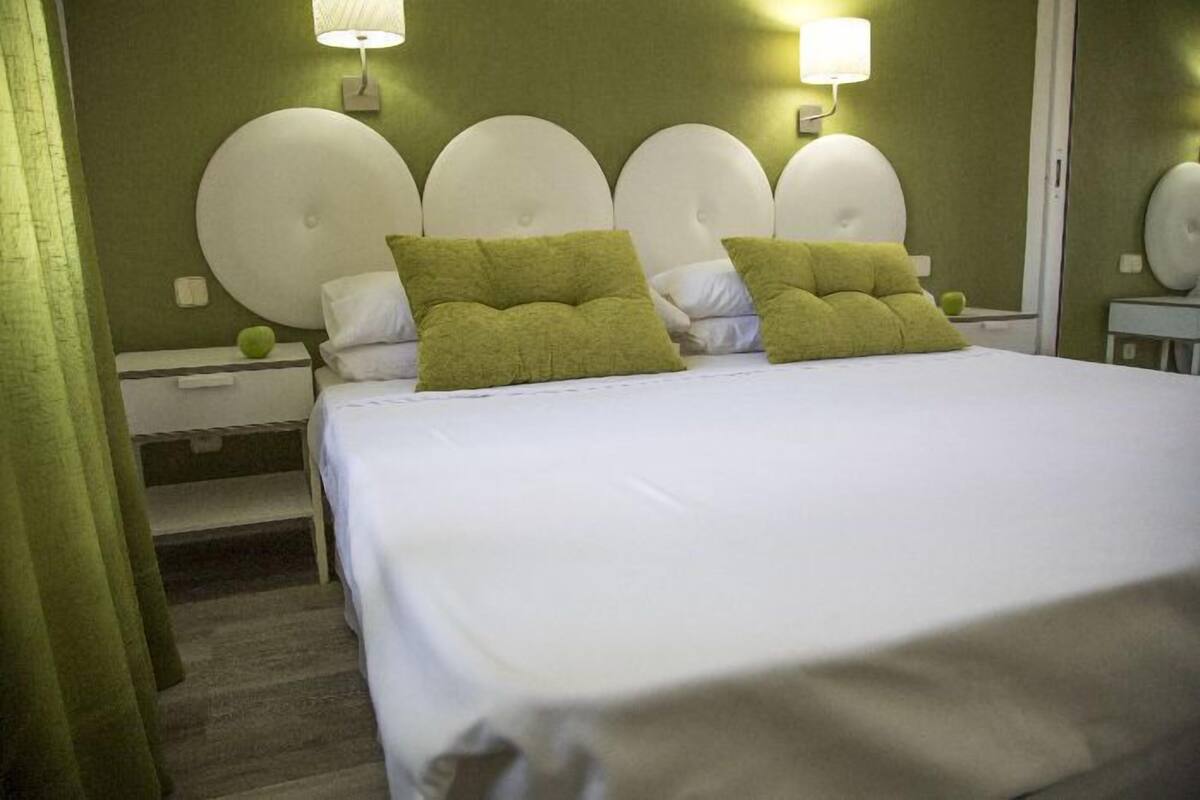 Une chambre d’hôtel avec un grand lit, deux oreillers verts, une tête de lit ronde et deux lampes fixées au mur.