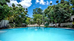 Piscina azul y cristalina rodeada de exuberante vegetación y árboles, con sillas de descanso y sombrillas cerca.