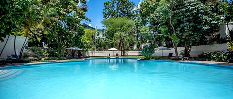 A clear blue swimming pool surrounded by lush greenery and trees, with lounge chairs and umbrellas nearby.