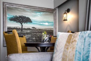 A room with a dining table, two chairs, a framed picture of a landscape, and a wall-mounted light fixture.