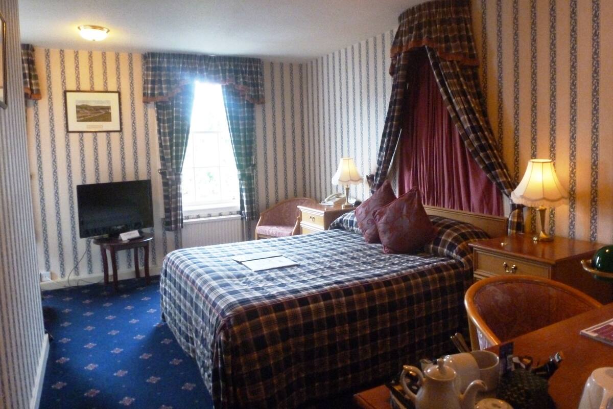 A hotel room with a bed, a television, a desk with a lamp, a chair, and a window with curtains.