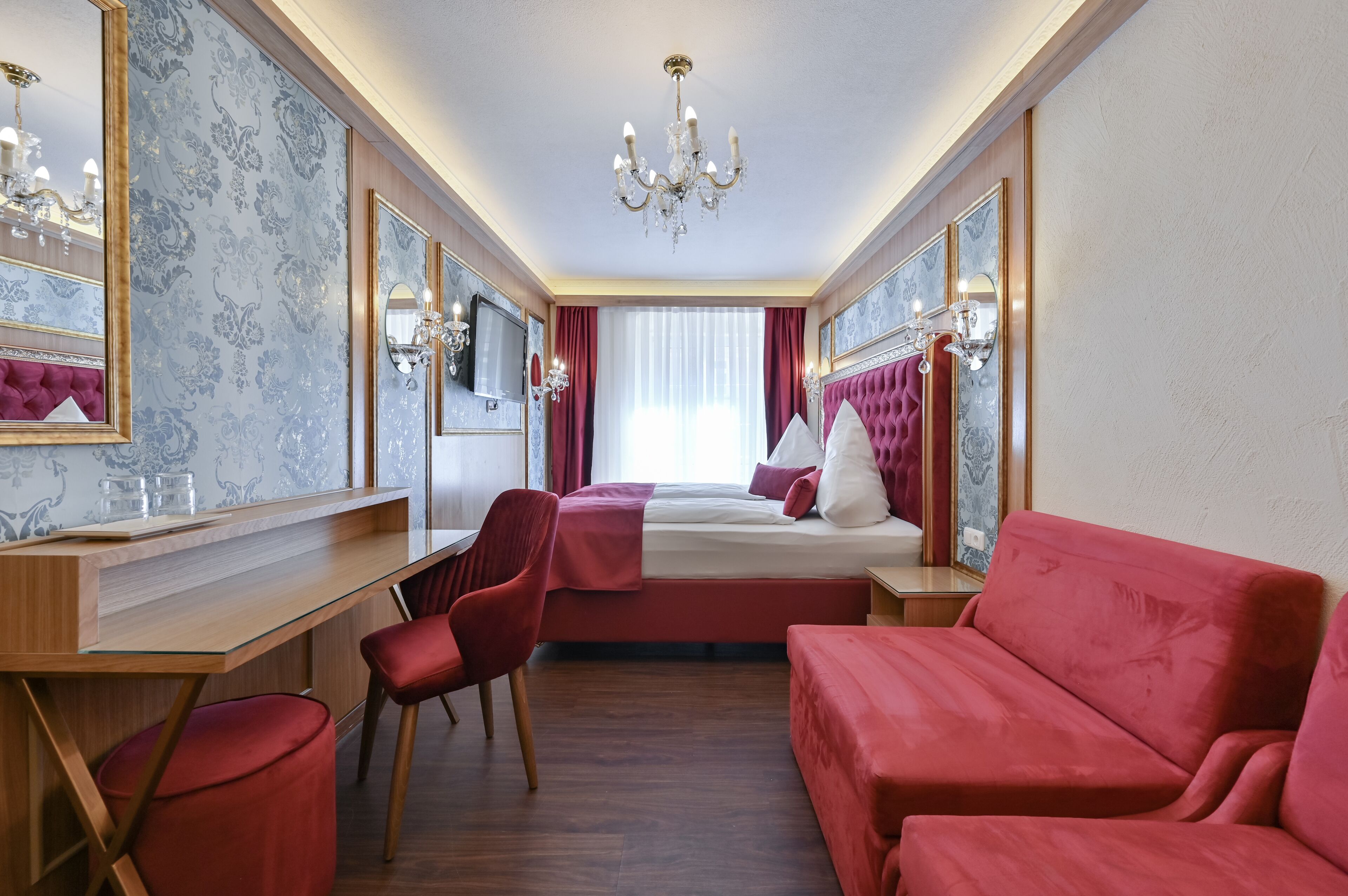 A hotel room with a bed, a desk, a chair, a sofa, and a chandelier.