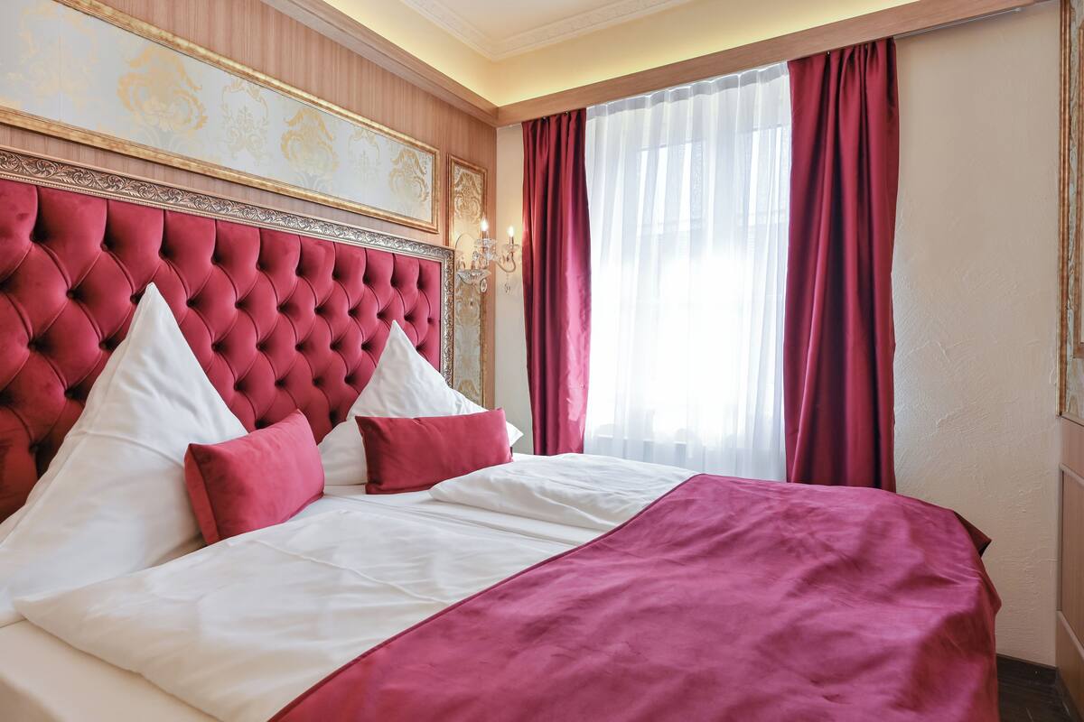 A hotel room with a tufted headboard, a large bed with white and red bedding, and a window with red curtains.