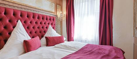 A hotel room with a tufted headboard, a large bed with white and red bedding, and a window with red curtains.
