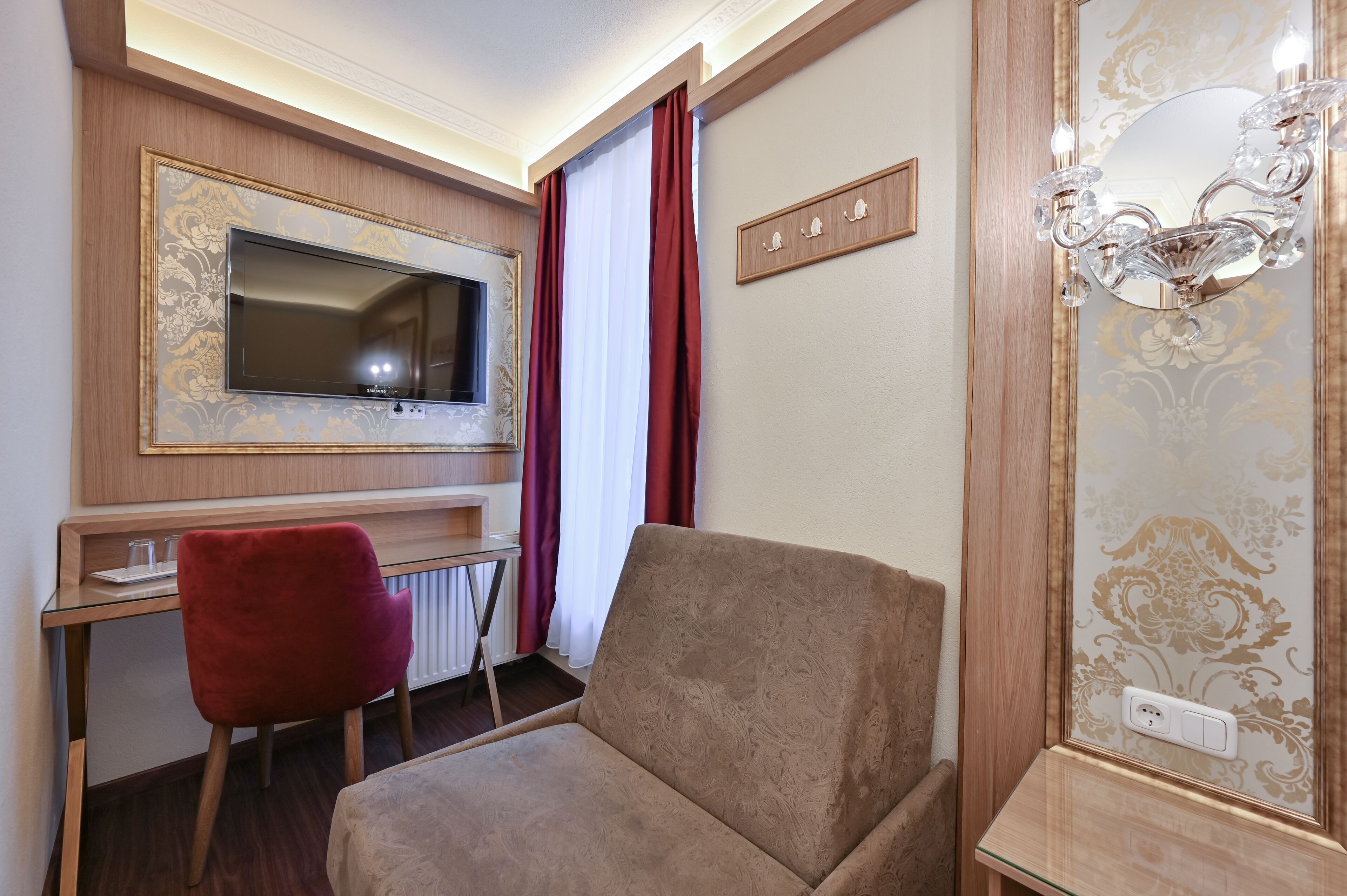 Camera d'albergo con televisore montato su un pannello di legno, una poltrona rossa, un divano beige e un lampadario.