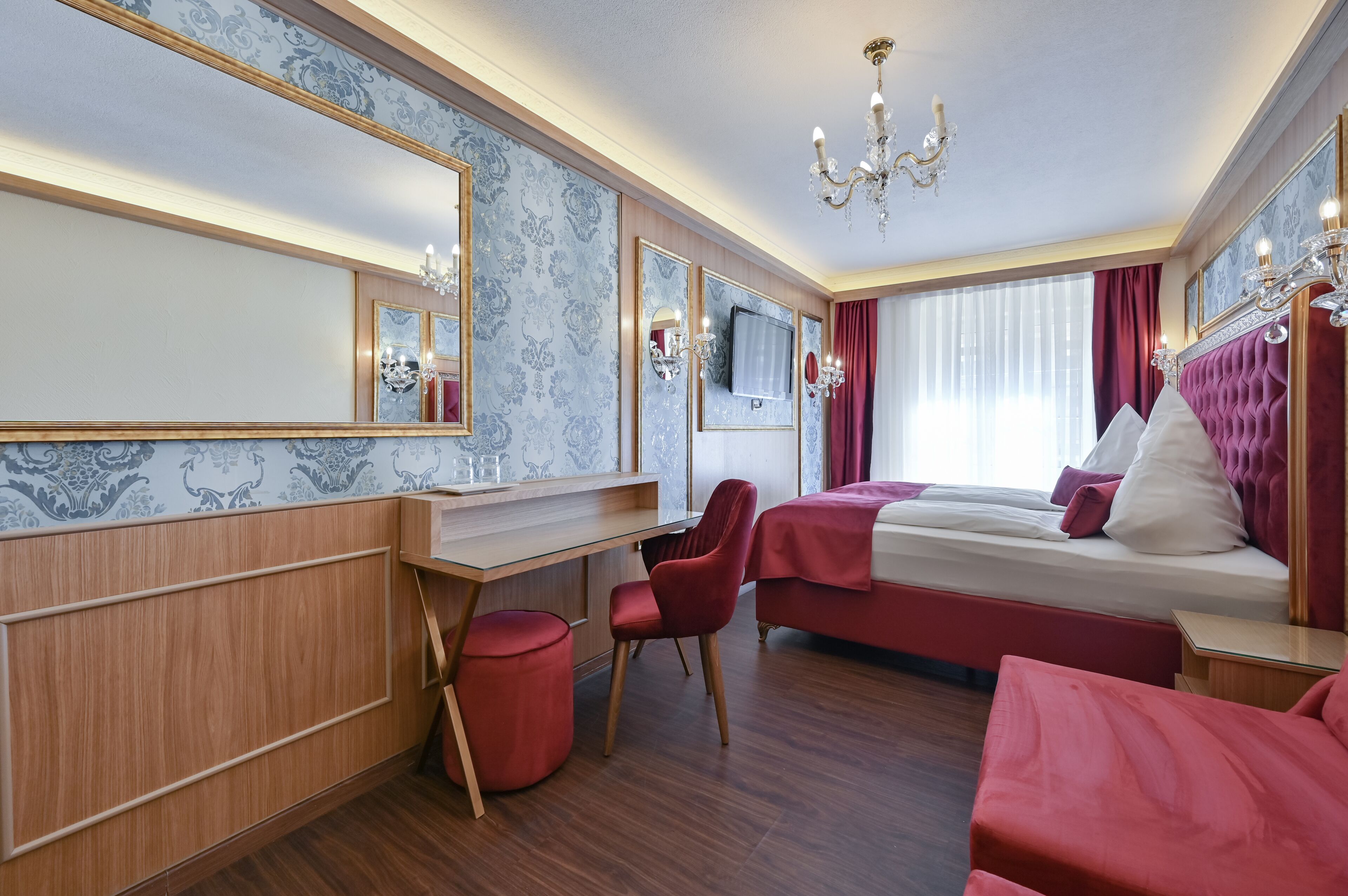 Een hotelkamer met een houten bureau, een rode stoel, een bed met rode beddengoed, een grote spiegel en een kroonluchter.