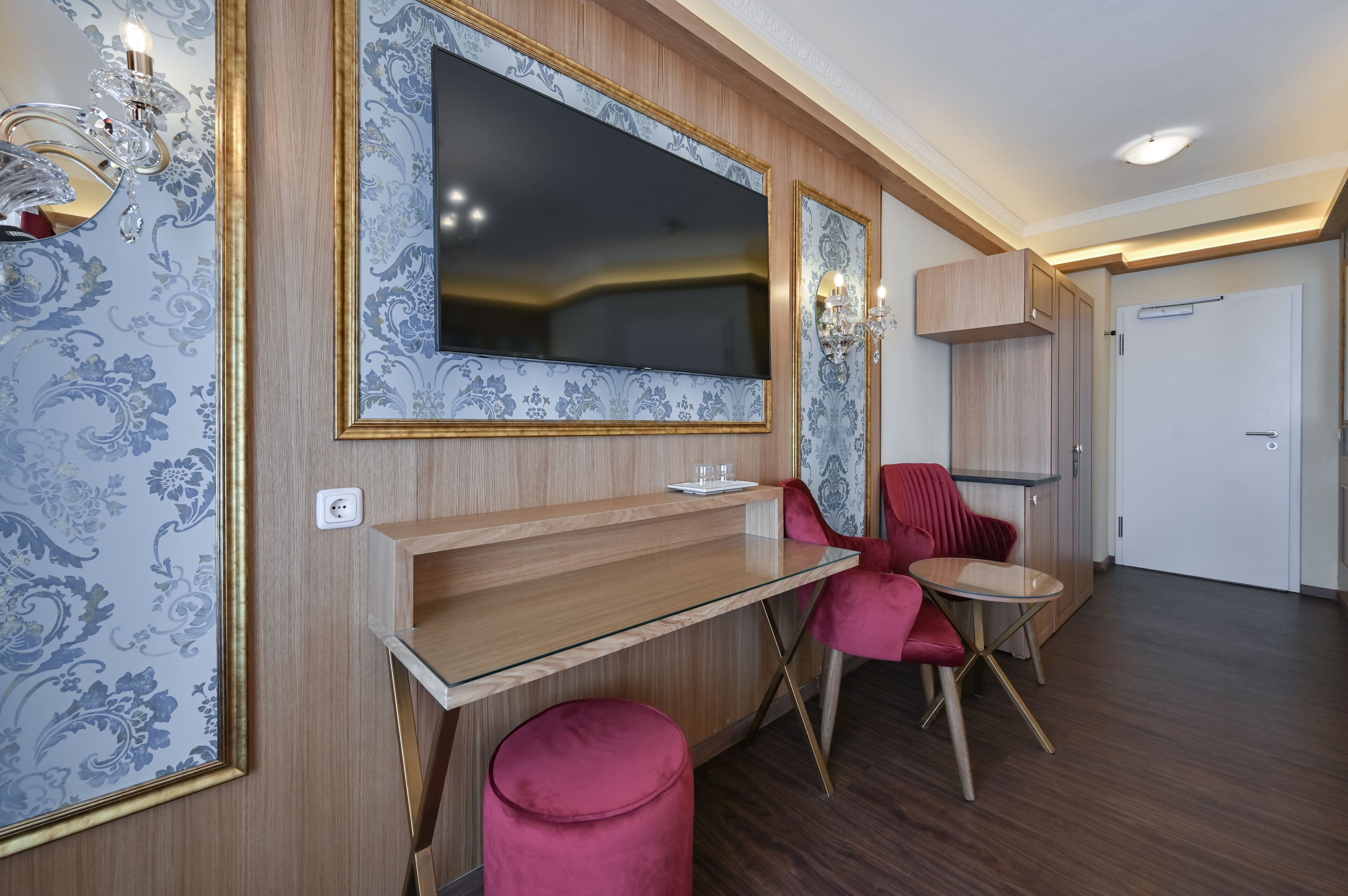 Una habitación moderna con un televisor de pantalla plana grande montado en una pared con papel tapiz decorativo, un escritorio de madera, una silla roja y un pequeño otoman.