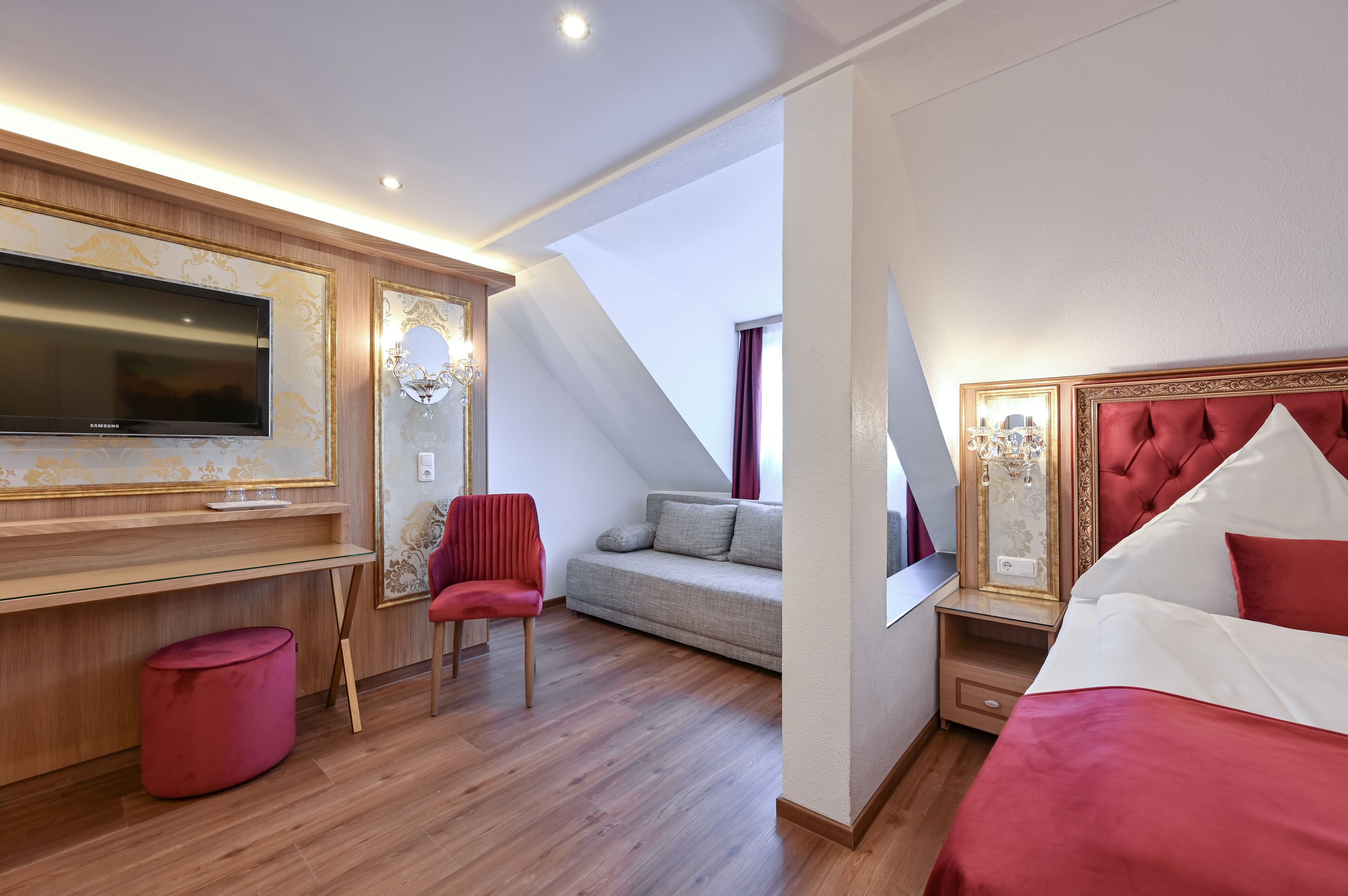 Una habitación de hotel moderna con una televisión de pantalla plana grande, un sillón tapizado en rojo, un sofá y una cama con ropa de cama roja.