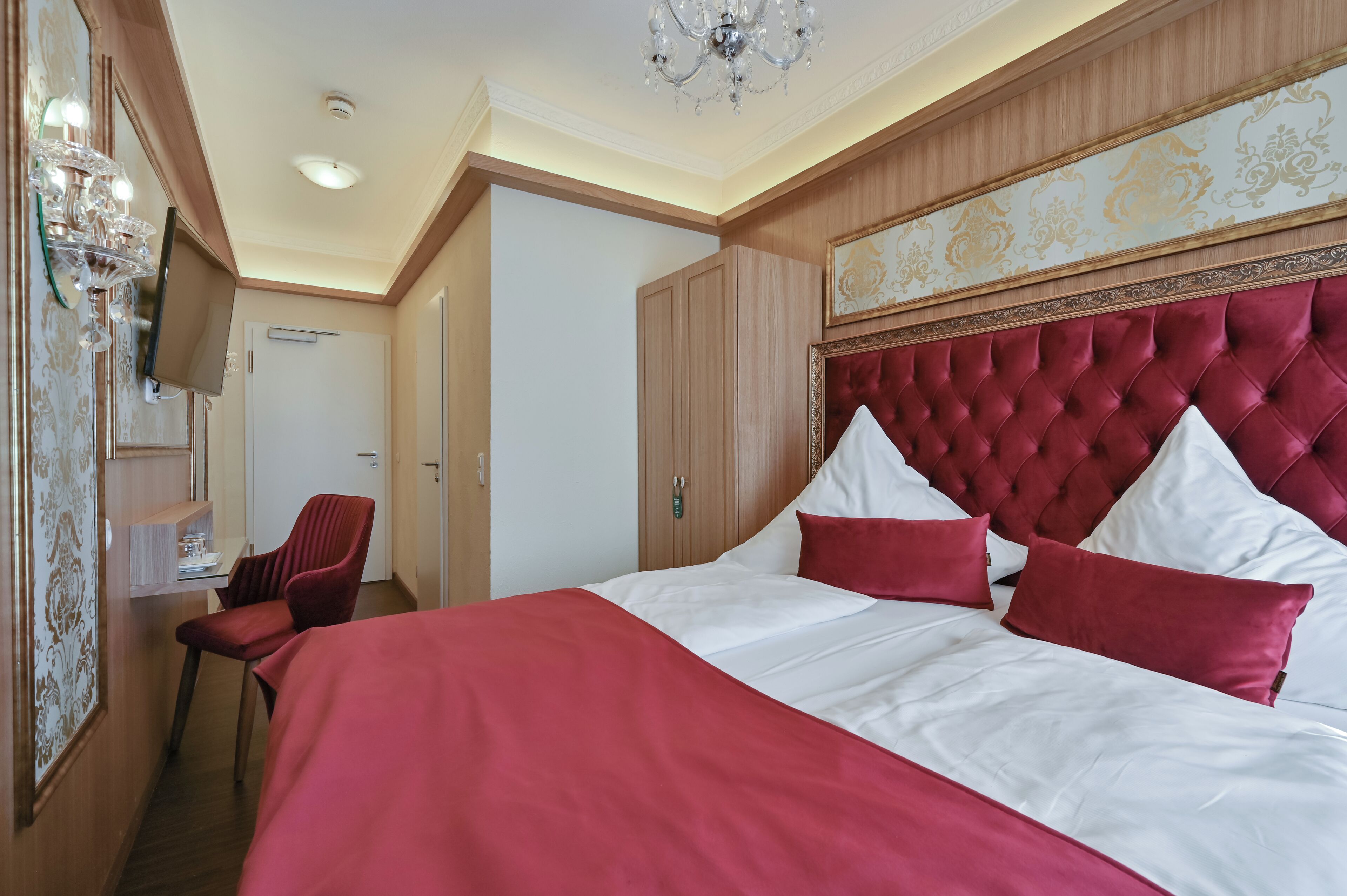 A hotel room with a large bed, a red chair, a television, and a chandelier.