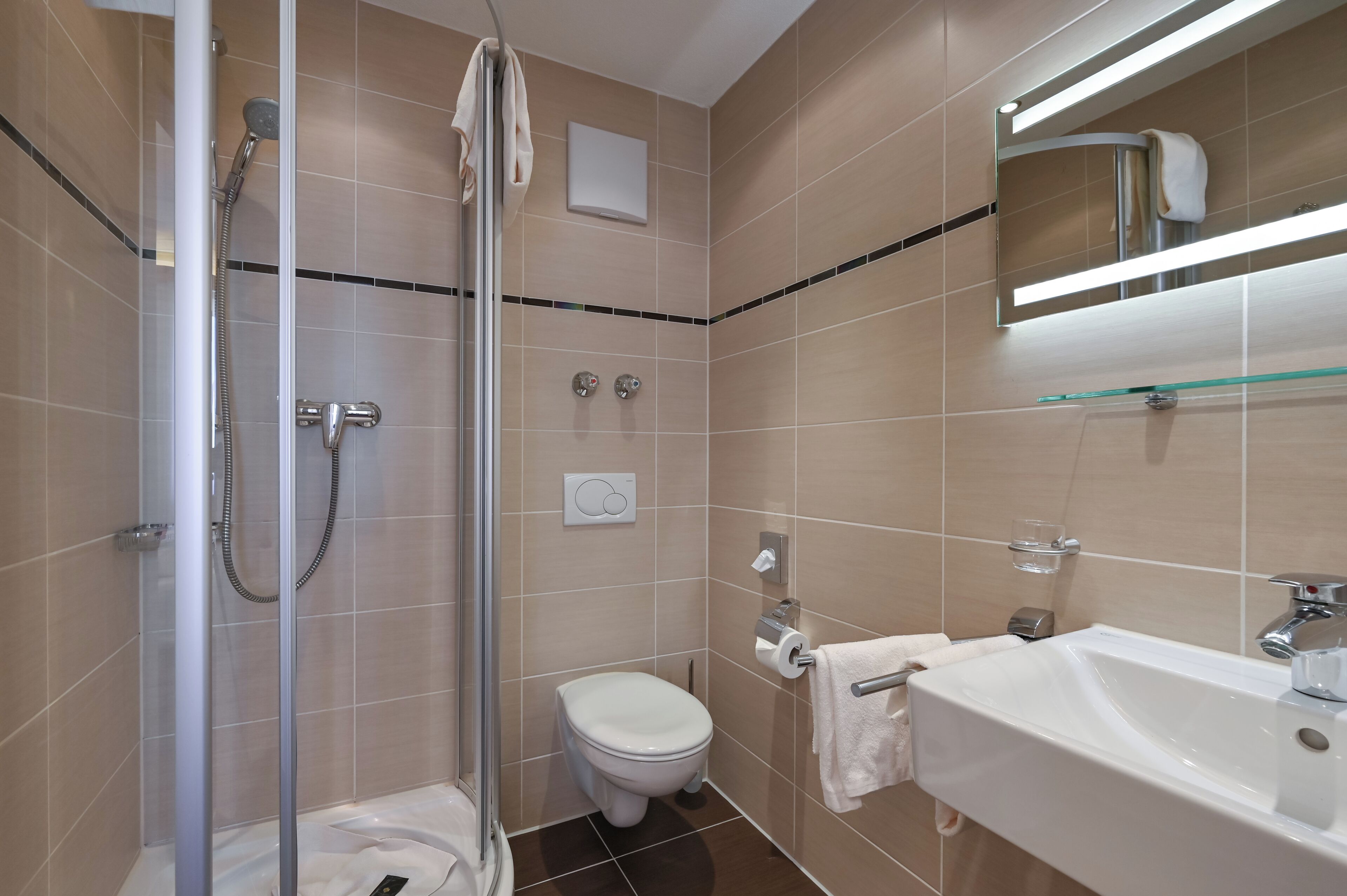 Un baño moderno con cabina de ducha de vidrio, inodoro blanco, lavamanos con espejo y paredes de azulejos color beige.