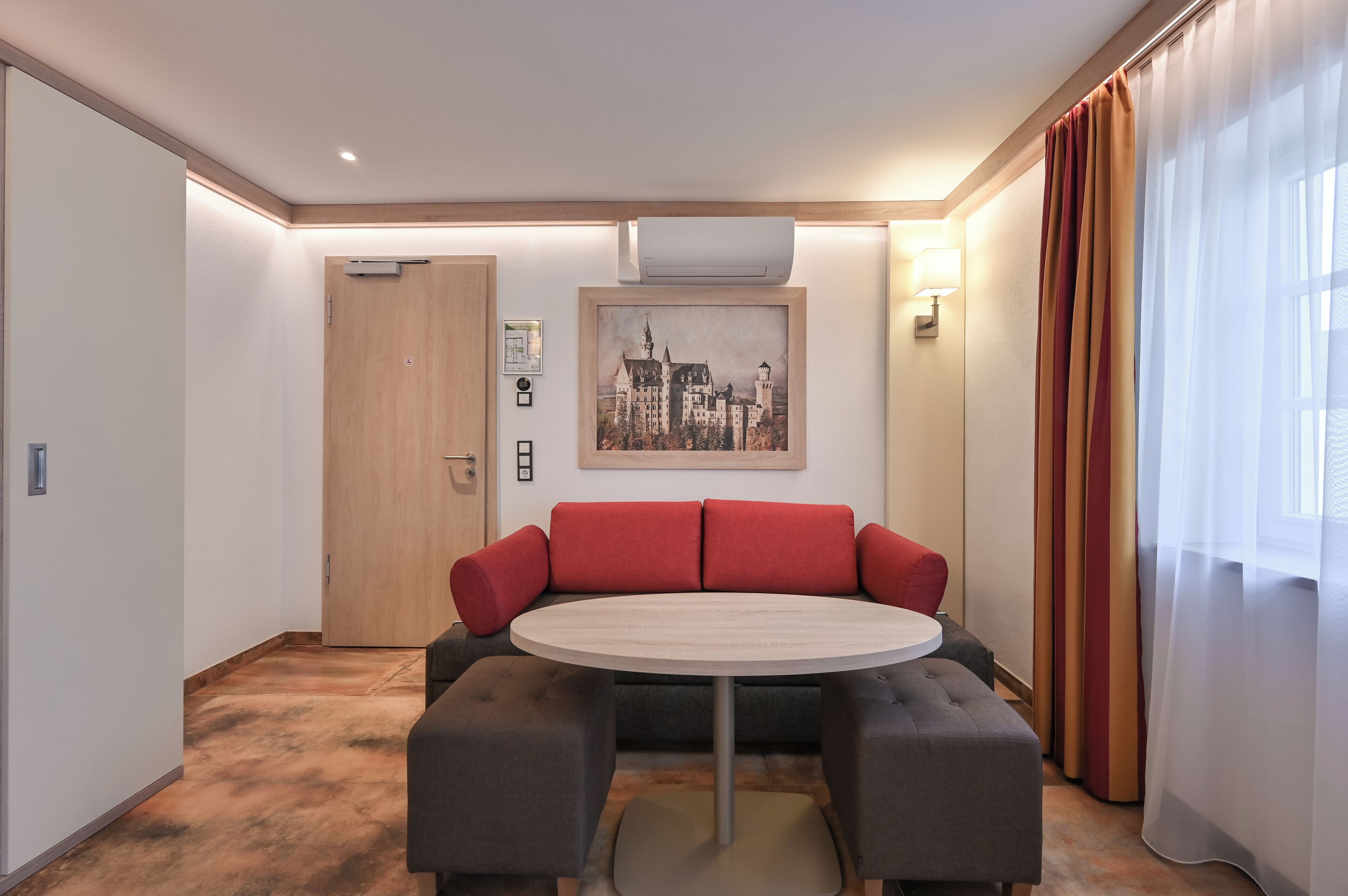 Une chambre d’hôtel avec une table ronde, un canapé rouge et gris, un tableau encadré représentant une vue sur la ville, et une porte en bois.