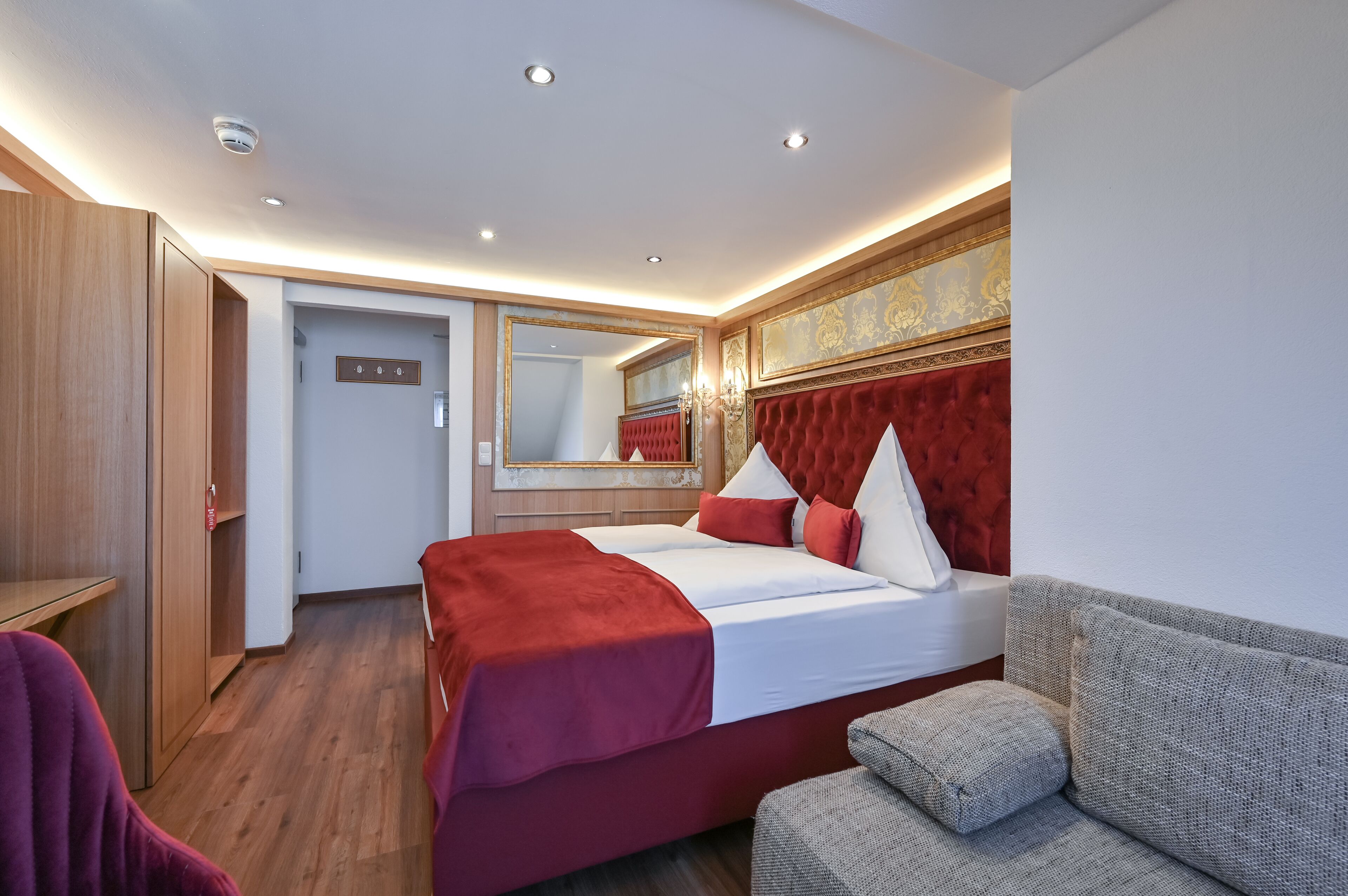 A hotel room with a large bed, a red headboard, a grey sofa, and a wooden wardrobe.