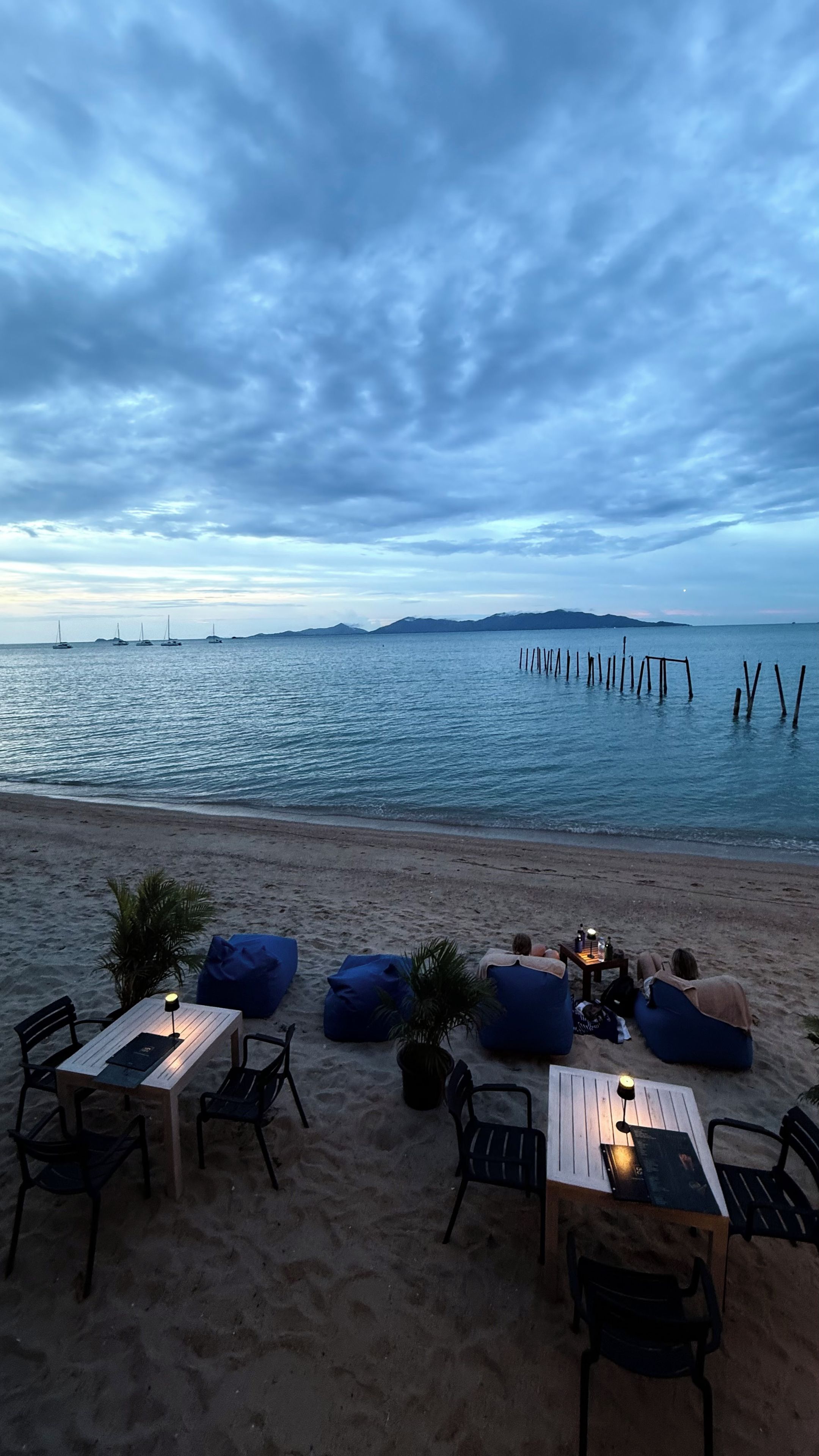 A beachside dining area with tables and chairs, a small table with a lamp, and a view of the sea with boats and a distant island.