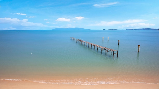 Una playa con un muelle de madera que se adentra en aguas tranquilas, rodeada de cielos despejados.
