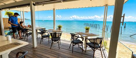 Terraza exterior con piso de madera, mesas y sillas, con vista a una playa y el mar.