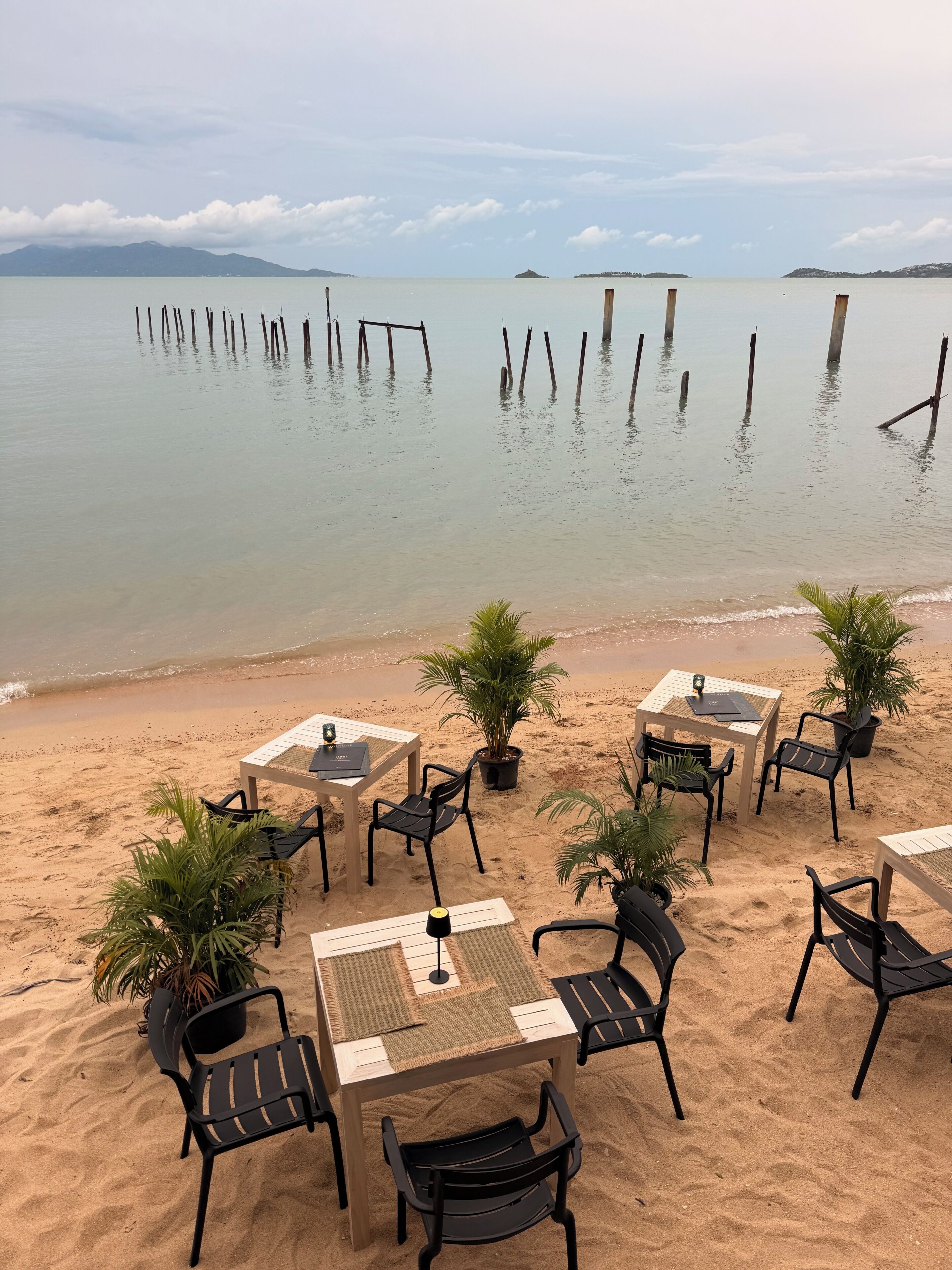 A beachside seating area with tables and chairs, surrounded by sand and overlooking calm water with wooden posts.