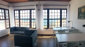 A room with a sofa, a dining table, and chairs, large windows with a view of the sea, and a painting on the wall. - Hotel Residencias Varadouro (Horta)