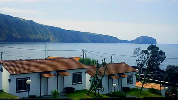 A coastal area with white buildings with red roofs, a view of the sea, and a hilly landscape in the background. - Hotel Residencias Varadouro (Horta)