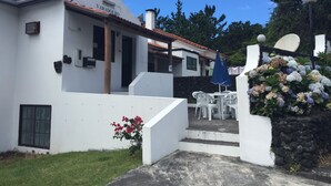 A white building with a tiled roof, a small garden with red flowers, and a white outdoor table with chairs. - Hotel Residencias Varadouro (Horta)