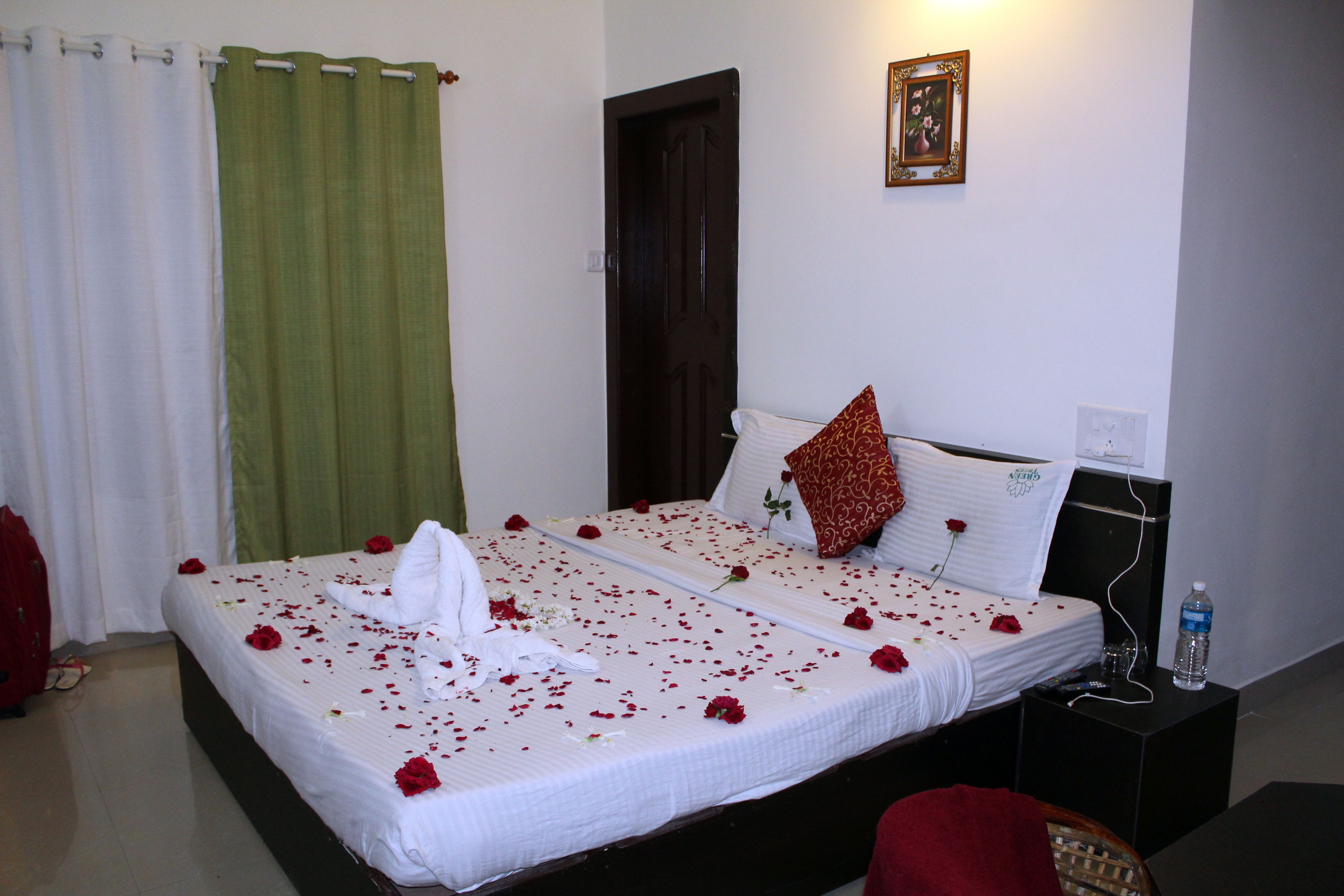 A neatly made bed with rose petals and a white towel, a green curtain, a wooden door, and a small bedside table with a water bottle.
