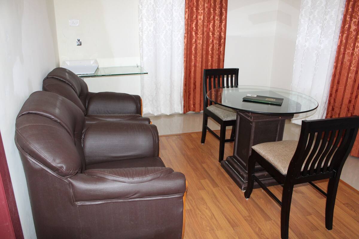 Una sala de estar con un sofá de cuero marrón, una mesa de comedor con superficie de vidrio, dos sillas de madera y un mueble blanco pequeño.