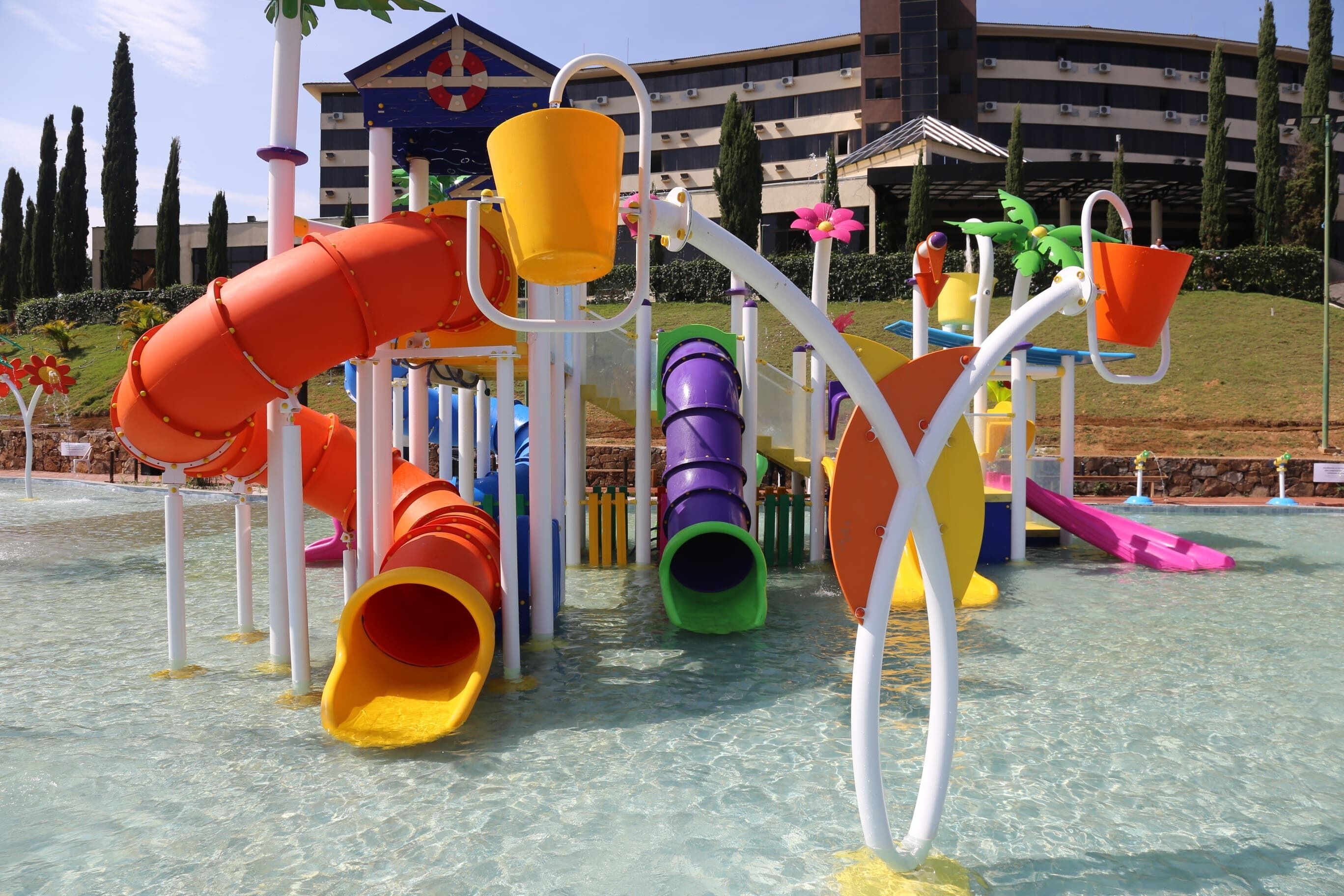 Un espace de jeux aquatiques doté de plusieurs toboggans et d’autres éléments aquatiques, entouré d’un bâtiment abritant une piscine et de la végétation.