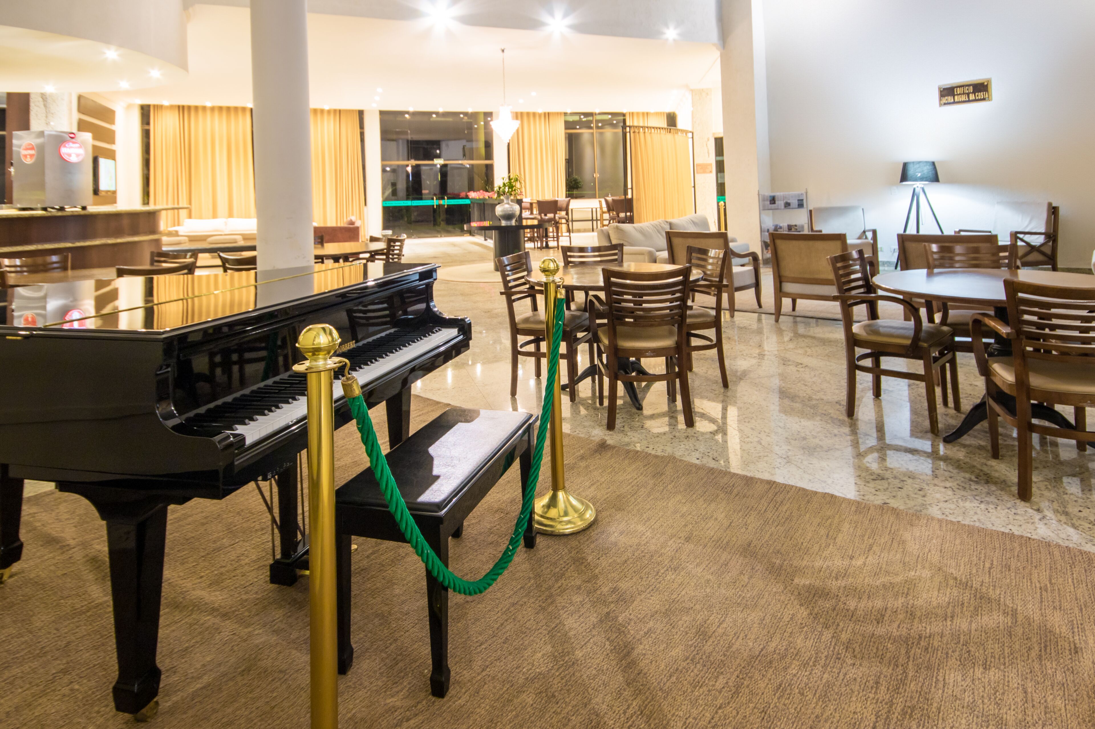 Um saguão de hotel com um piano de cauda, área de estar com mesas e cadeiras, e uma grande janela com cortinas.