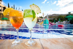 Twee cocktails op een betegelde tafel bij het zwembad, met zwembad en parasols op de achtergrond.