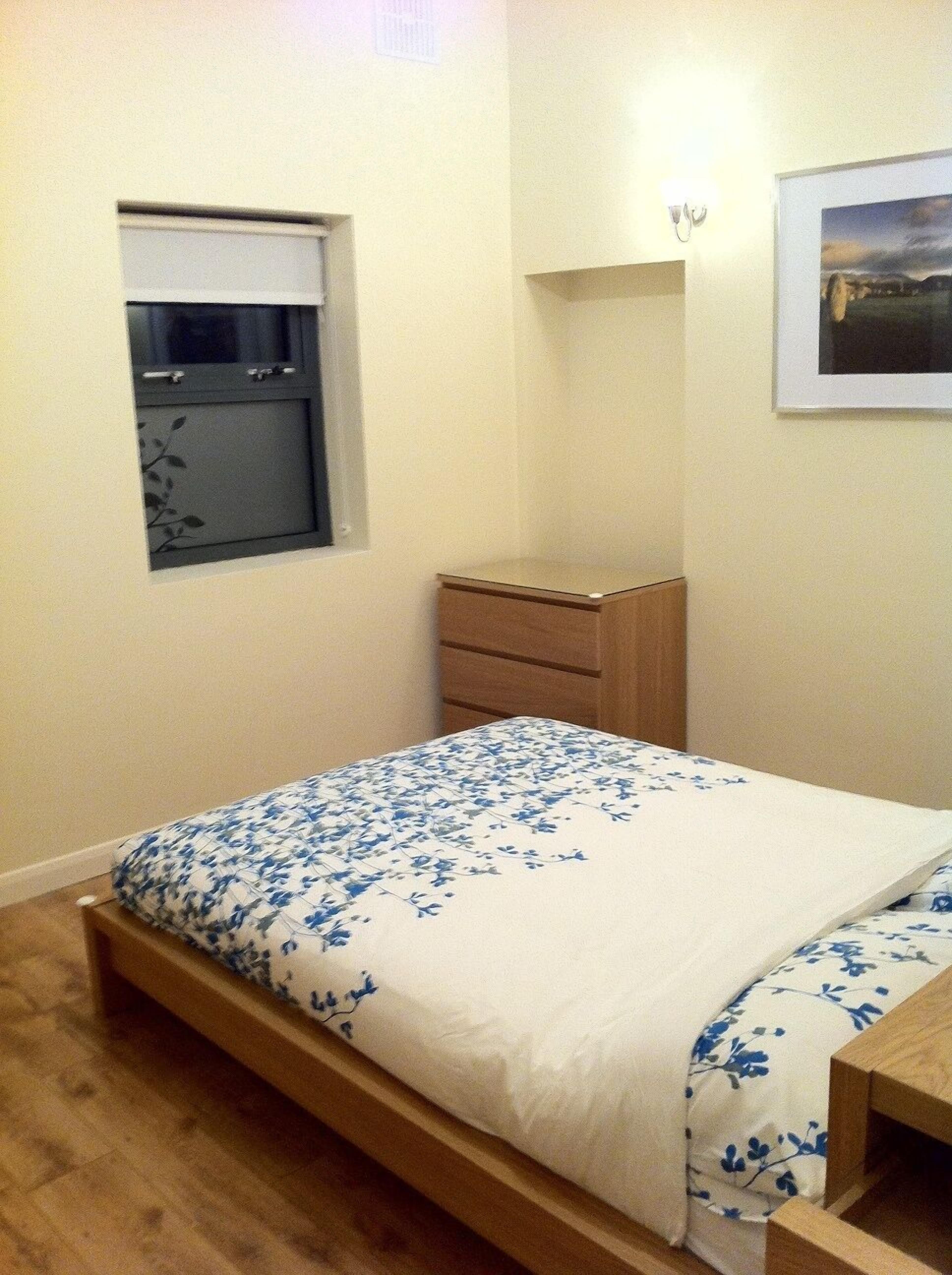 A bedroom with a single bed, a wooden nightstand, a small dresser, a window with a closed blind, and a framed picture on the wall.