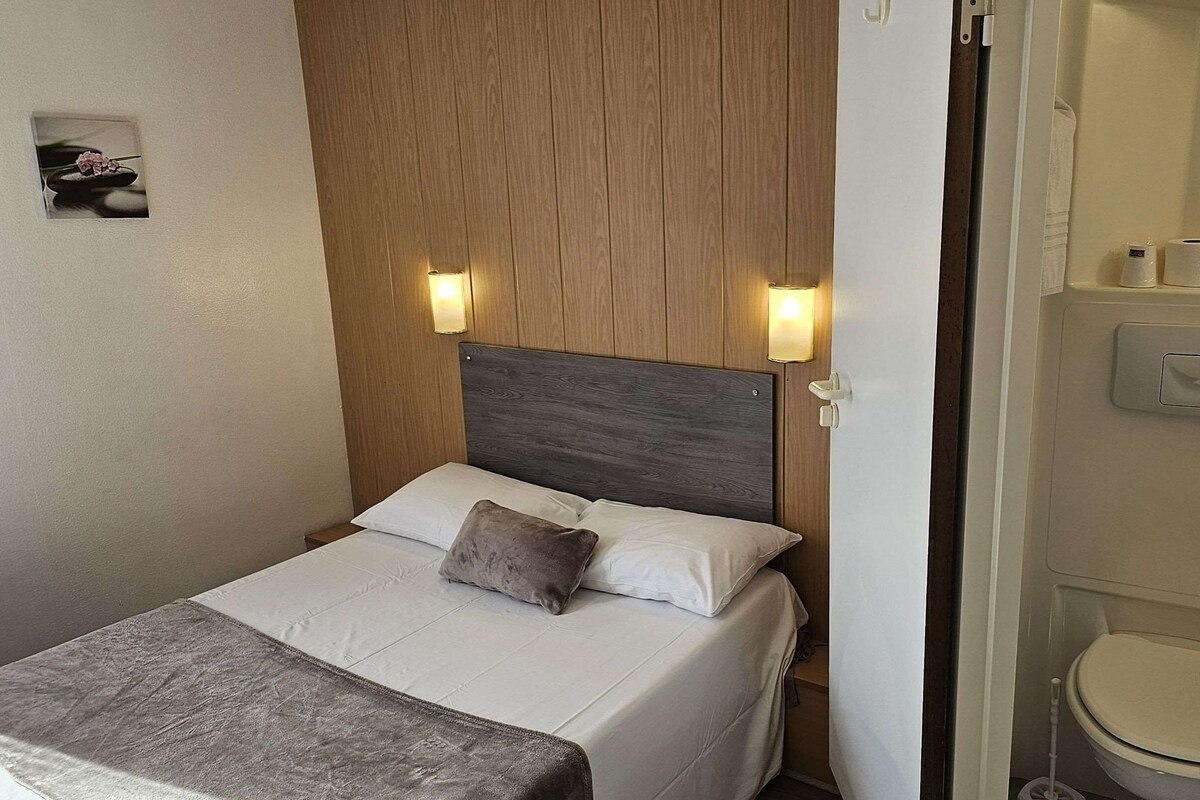 A hotel room with a bed, a wooden headboard, two wall-mounted lights, a small picture on the wall, and a bathroom with a toilet and a hand dryer.