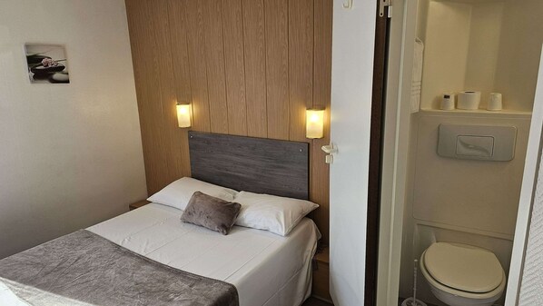 A hotel room with a bed, a wooden headboard, two wall-mounted lights, a small picture on the wall, and a bathroom with a toilet and a hand dryer.