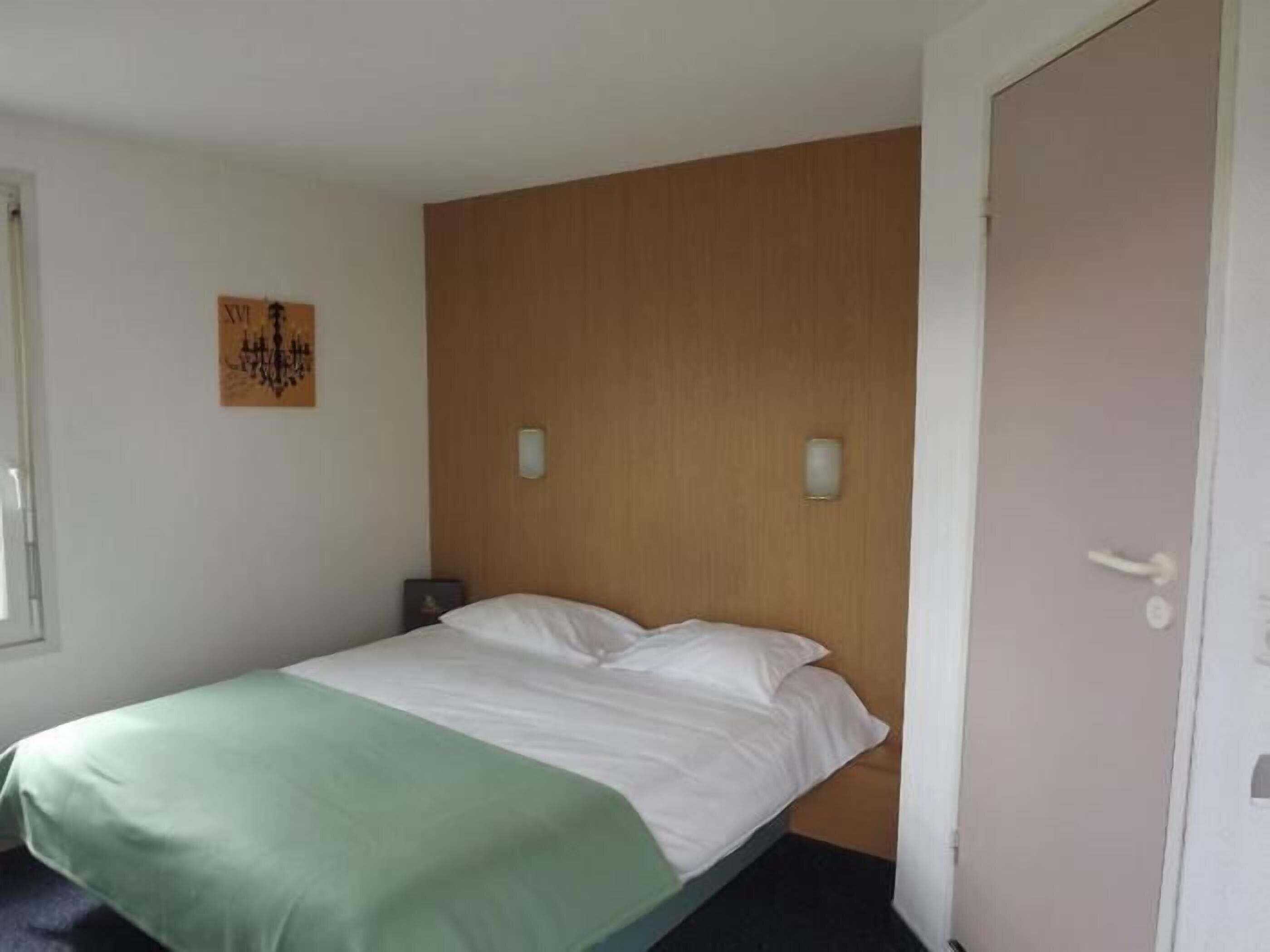 A single bed with a green and white bedspread, a wooden headboard, two wall-mounted light fixtures, and a door with a handle.