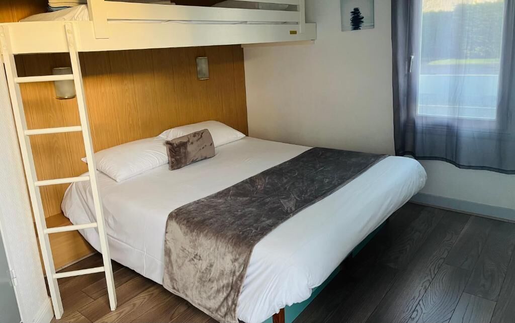 A bunk bed room with a ladder, a wooden headboard, a bedside table, and a window with a sheer curtain.