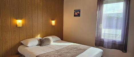 Una cama individual con cabecera de madera, dos lámparas fijadas a la pared, una ventana con cortina y un pequeño cuadro floral.