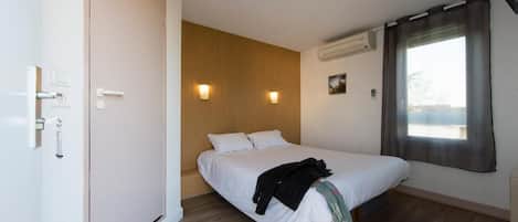 A bedroom with a bed, wooden panel wall, two wall-mounted lights, an air conditioner, and a window with curtains.