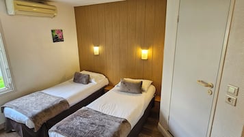 Et hotelværelse med to enkeltsenge, træpaneler på væggen, to vægmonterede lamper og en dør med håndtag og lås.